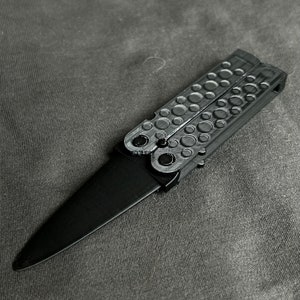 May include: A black butterfly knife with a silver handle. The handle has a textured design with small circles.