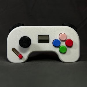 May include: A white plastic video game controller with black buttons and a red, blue, green, and pink button on the front.