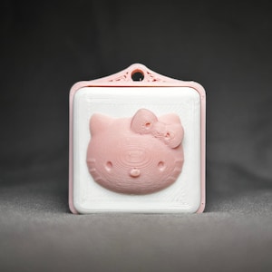 May include: A pink plastic keychain with a white square frame featuring a 3D embossed image of Hello Kitty.
