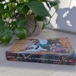 May include: A hardcover book titled Harry Potter and the Prisoner of Azkaban by Rowling, featuring a cover and spine completely covered in small, multicolored, reflective rhinestones that create a textured, sparkling mosaic effect across the surface.