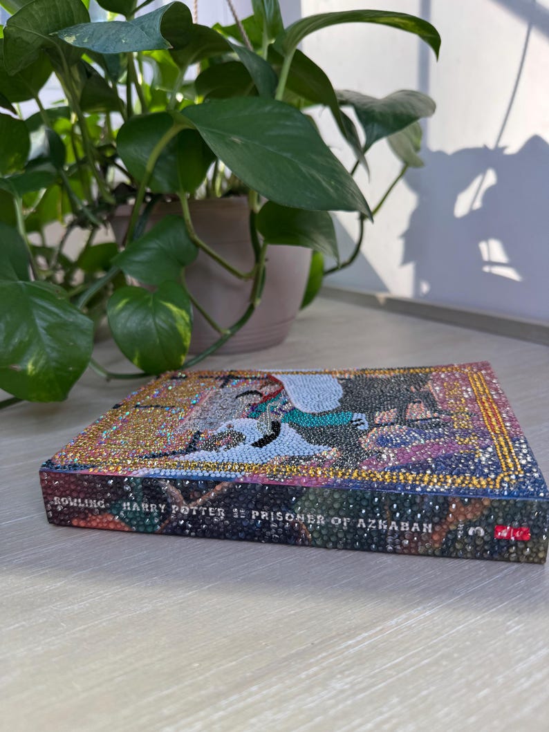 May include: A hardcover book titled Harry Potter and the Prisoner of Azkaban by Rowling, featuring a cover and spine completely covered in small, multicolored, reflective rhinestones that create a textured, sparkling mosaic effect across the surface.