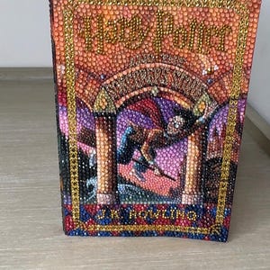 May include: A book cover featuring a diamond art mosaic of Harry Potter and the Sorcerer's Stone by J.K. Rowling. The design is composed of small, colorful, reflective rhinestones in shades of orange, purple, blue, and gold, forming the title and illustration.