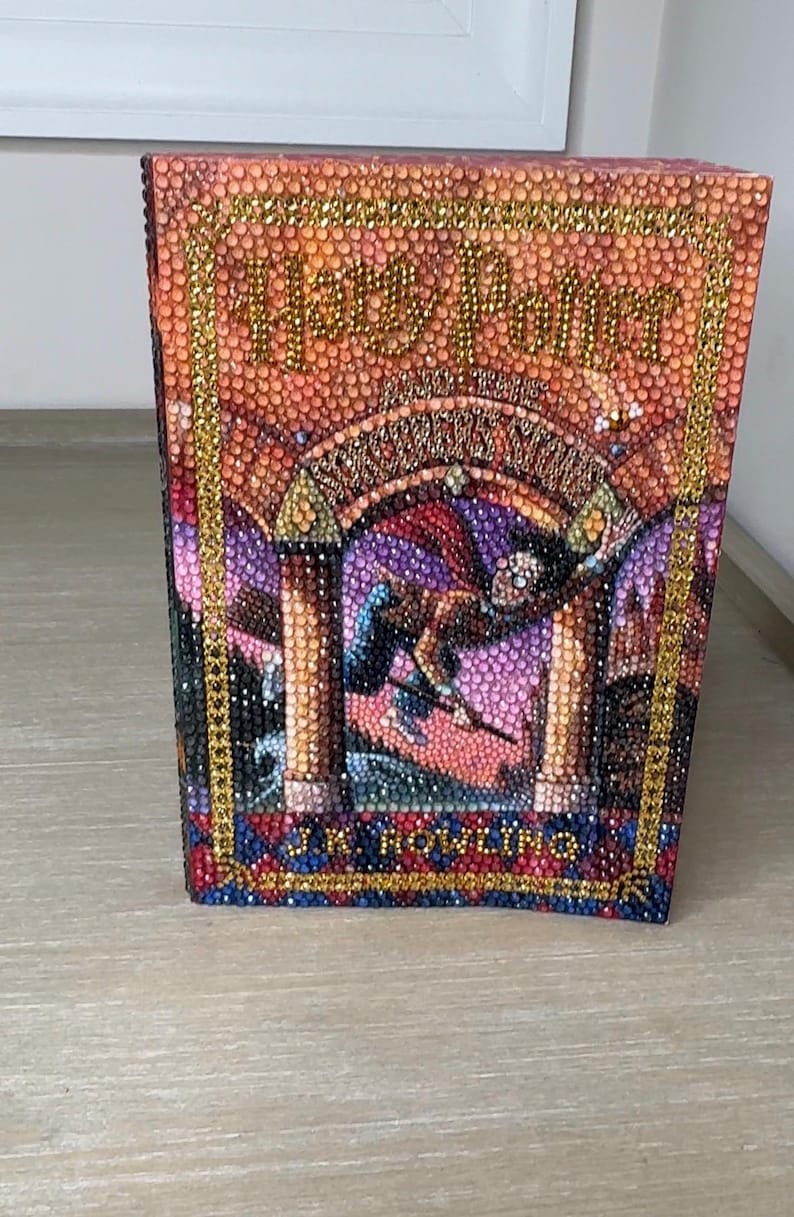 May include: A book cover featuring a diamond art mosaic of Harry Potter and the Sorcerer's Stone by J.K. Rowling. The design is composed of small, colorful, reflective rhinestones in shades of orange, purple, blue, and gold, forming the title and illustration.