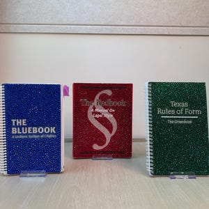 May include: Three spiral-bound books covered in sparkling rhinestones. The blue book reads THE BLUEBOOK A Uniform System of Citation. The red book features a section symbol and reads The Redbook A Manual On Legal Style. The green book reads Texas Rules of Form The Greenbook.