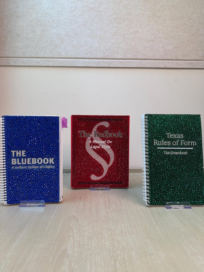 May include: Three spiral-bound books covered in sparkling rhinestones. The blue book reads THE BLUEBOOK A Uniform System of Citation. The red book features a section symbol and reads The Redbook A Manual On Legal Style. The green book reads Texas Rules of Form The Greenbook.