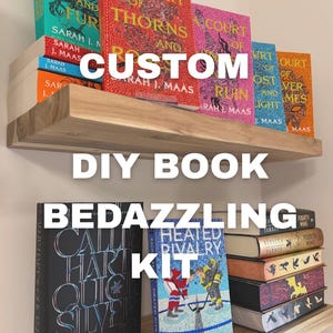 Può includere: Un kit fai-da-te per decorare libri esposto su due mensole in legno. Le mensole contengono vari libri con copertine colorate, inclusi titoli di Sarah J. Maas e Rachel Reid, caratterizzati da perline intricate e decorazioni applicate.