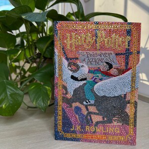 May include: A hardcover book titled Harry Potter and the Prisoner of Azkaban by J.K. Rowling, featuring a cover entirely embellished with small, colorful, reflective rhinestones. The design depicts a winged creature and text in gold, pink, and blue.