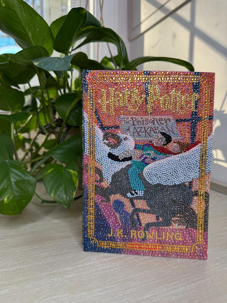 May include: A hardcover book titled Harry Potter and the Prisoner of Azkaban by J.K. Rowling, featuring a cover entirely embellished with small, colorful, reflective rhinestones. The design depicts a winged creature and text in gold, pink, and blue.