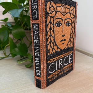 May include: A book titled Circe by Madeline Miller, featuring a cover and spine completely covered in black and orange rhinestones. The design includes a stylized face illustration and geometric border patterns, with the title and author text clearly visible.