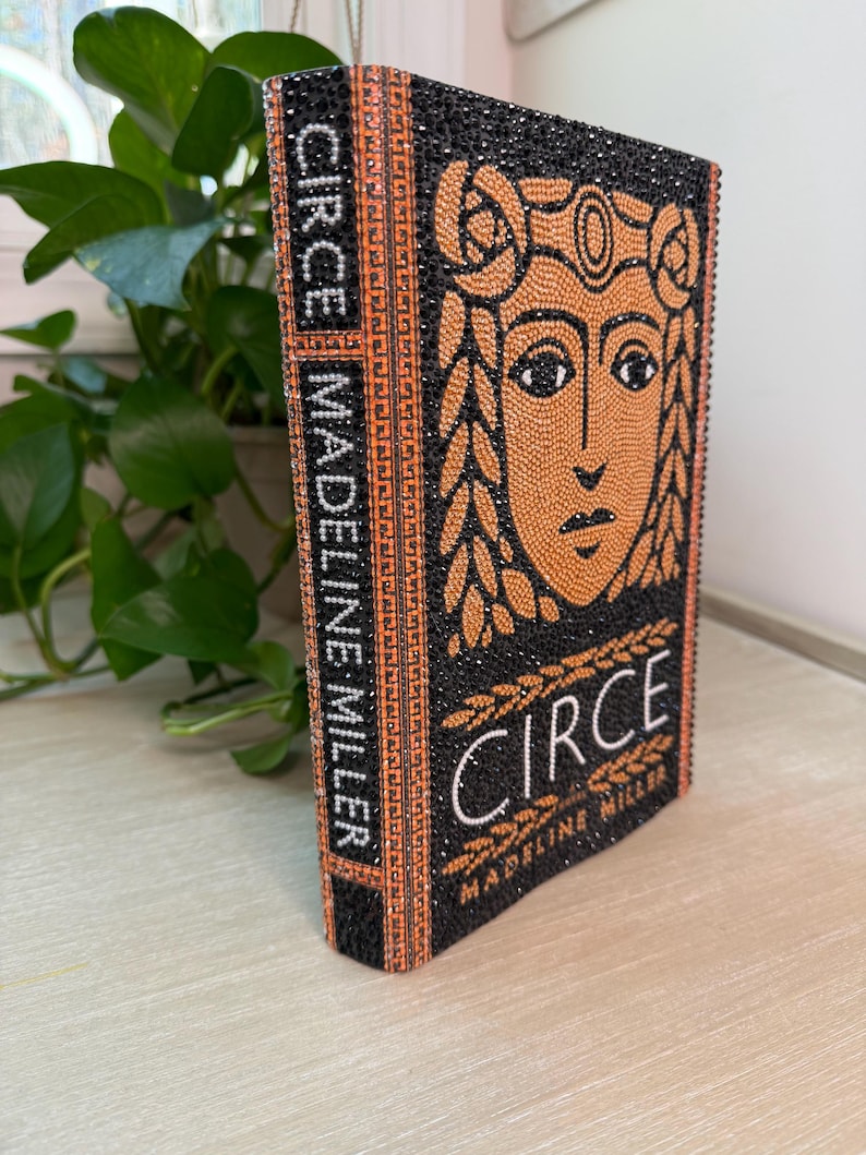 May include: A book titled Circe by Madeline Miller, featuring a cover and spine completely covered in black and orange rhinestones. The design includes a stylized face illustration and geometric border patterns, with the title and author text clearly visible.