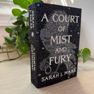 May include: A black book titled "A Court of Mist and Fury" by Sarah J. Maas. The cover is adorned with black crystals and iridescent gems. The title and author's name are in silver lettering.