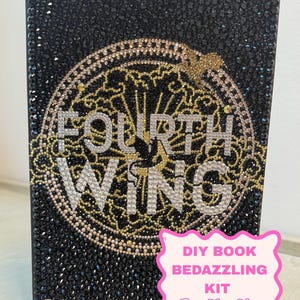 May include: A book cover decorated with black, gold, and clear rhinestones. The text "FOURTH WING" and "REBECCA" are spelled out in crystals. A circular gold filigree design surrounds the title. A pink sign reads "DIY BOOK BEDAZZLING KIT".