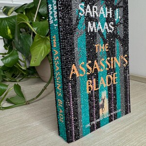 May include: A hardcover book titled The Assassin's Blade by Sarah J. Maas, featuring a cover entirely embellished with small black, teal, silver, and orange rhinestones. The text and a small deer silhouette are formed by the colored stones.