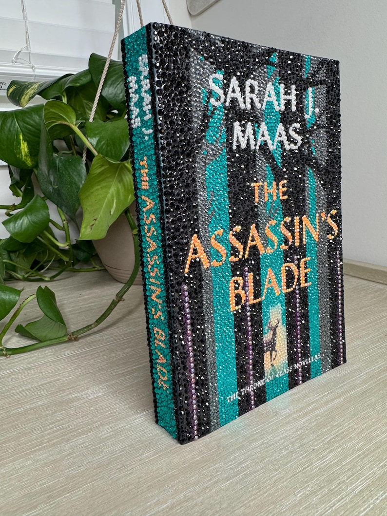 May include: A hardcover book titled The Assassin's Blade by Sarah J. Maas, featuring a cover entirely embellished with small black, teal, silver, and orange rhinestones. The text and a small deer silhouette are formed by the colored stones.