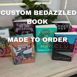 May include: Several custom bedazzled books are displayed, including titles such as "The Hunger Games" and "The Club." The books are adorned with sparkling rhinestones, and the text "CUSTOM BEDAZZLED BOOK MADE TO ORDER" is visible.