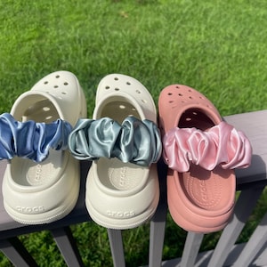 May include: Three pairs of white clogs with different colored satin scrunchies around the ankle strap. The scrunchies are blue, green, and pink.