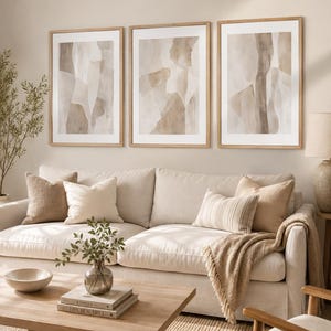 May include: A set of three framed vertical wall art prints featuring abstract watercolor shapes in shades of beige, cream, and taupe. Each print is encased in a light-toned wooden frame with a white mat, displayed in a horizontal row on a wall.