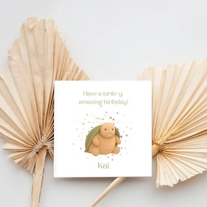 May include: A square white greeting card featuring a cartoon turtle illustration in the centre with the text "Have a turtle-y amazing birthday!" above it and the name "Kai" printed in a simple green font below the turtle graphic.