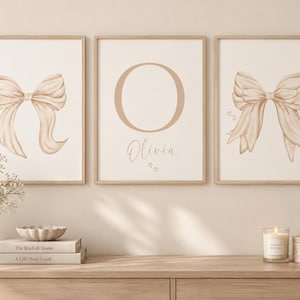 Op de afbeelding: Drie ingelijste wandprints met een beige strik aan de linkerkant, de letter O en de naam Olivia in het midden, en een beige strik met kleine hartjes aan de rechterkant. De prints zitten in lichte houten lijsten aan een muur.