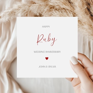 May include: A square white greeting card featuring the black text HAPPY, the red script text Ruby, the black text WEDDING ANNIVERSARY, a small red heart icon, and the black text JOHN & SYLVIA centred on the front of the card.