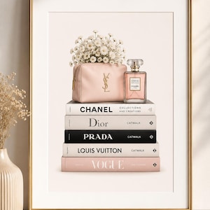 Puede incluir: Una lámina enmarcada con una pila de cinco libros de moda titulados Chanel, Dior, Prada, Louis Vuitton y Vogue. Sobre los libros hay un neceser rosa claro con un logotipo dorado y un frasco de perfume de cristal junto a flores blancas.