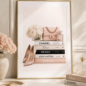Può includere: Una stampa incorniciata con una pila di libri di moda intitolati Chanel, Dior, Prada, Louis Vuitton e Vogue. Sopra i libri si trovano una borsa rosa trapuntata, un flacone di profumo, un bouquet di fiori bianchi e due décolleté beige.