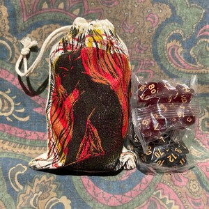 May include: A drawstring dice bag with a black and red illustration, next to a package of burgundy dice. The bag features a white drawstring and is set against a patterned fabric background. The dice are visible through clear packaging.