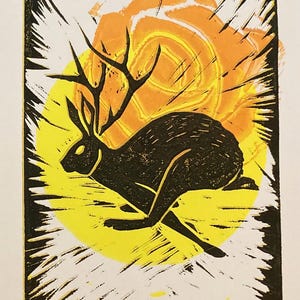Può includere: Una stampa a linoleum in nero e giallo di un coniglio che corre con le corna, su uno sfondo di sole giallo e arancione.