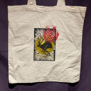 Può includere: Borsa tote bianca con una grafica nera e gialla di una lepre che corre con le corna, su uno sfondo di sole giallo e rosa rossa. La borsa ha due manici ed è realizzata in tessuto strutturato.