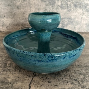 May include: A teal ceramic serving bowl with a raised center. The bowl has a wide, shallow base and a smaller, cup-like structure rising from the center. The glaze has a speckled appearance, with darker blue accents around the rim.