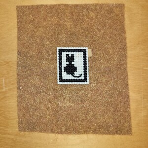 May include: A small, square cross-stitch featuring a black cat silhouette within a white and black border. The needlework is centered on a brown felt square, with a small tan tag attached.