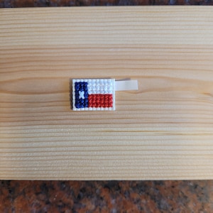 May include: A small, hand-stitched Texas flag patch with a white, blue, and red design. The patch is attached to a piece of wood with a white adhesive strip. The flag features a white star on a blue background, with red and white stripes.
