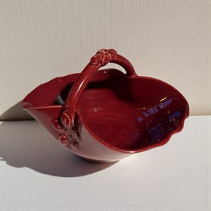 May include: A burgundy ceramic basket with a handle. The basket has a scalloped edge and a floral design on the side. The handle is also decorated with floral details. The basket is likely for decorative use.