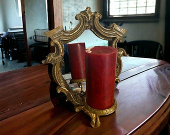 Könnte beinhalten: Ein goldfarbener Metallkerzenhalter mit einem verzierten, geschwungenen Rahmen und integriertem Spiegel. Eine einzelne, dicke, zylindrische rote Stumpenkerze steht auf dem runden Sockel und spiegelt sich im dahinterliegenden Spiegel.
