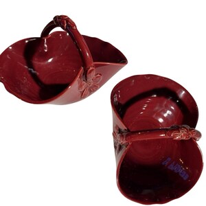 May include: Two burgundy ceramic baskets with handles. One basket is in a side view, the other is a top view. Both have floral accents on the sides and handle attachments. The baskets are likely for decorative or serving purposes.