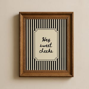 May include: A framed print with a brown wooden frame. The print features a black and white striped background with a cream-colored central panel. The panel has the words "Hey sweet cheeks" in black script.
