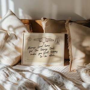 Puede incluir: Una postal vintage enmarcada con el texto "Always find time for the things that make you feel happy to be alive." La postal est&aacute; rodeada de almohadas beige y una manta blanca.