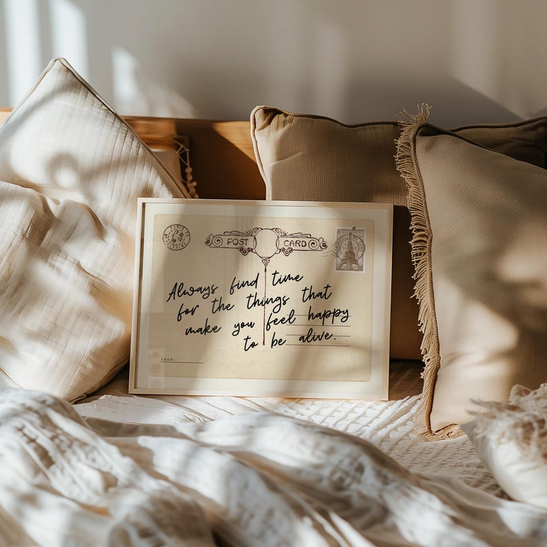 Puede incluir: Una postal vintage enmarcada con el texto "Always find time for the things that make you feel happy to be alive." La postal est&aacute; rodeada de almohadas beige y una manta blanca.