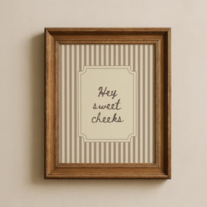 May include: A framed print with a brown wooden frame. The print has a beige background with vertical stripes in shades of beige and brown. The centre has a cream-coloured label with the words "Hey sweet cheeks" in a script font.