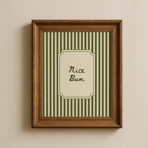 Op de afbeelding: Een ingelijste print met een bruine houten lijst. De print heeft een crèmekleurige achtergrond met groene verticale strepen. Het midden heeft een crèmekleurige rechthoek met de woorden "Nice Bum" in zwart schrift.