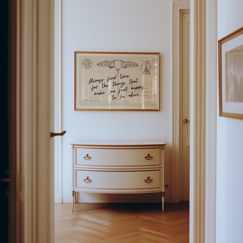 Puede incluir: Una impresi&oacute;n enmarcada de postal con el texto "Always find time for the things that make you feel happy to be alive" cuelga sobre una c&oacute;moda blanca y dorada con dos cajones. La habitaci&oacute;n tiene un ambiente luminoso y aireado.