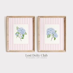 May include: Two framed prints featuring watercolor paintings of blue hydrangea flowers. The artwork is set against a white background with a pink striped border. The frames are a light brown color. The text "Lost Deity Club Printable Art Studio" is at the bottom.
