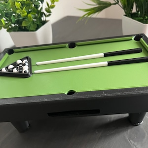 Può includere: Un biliardo verde in miniatura con finiture e gambe nere. Il biliardo ha un triangolo di deposito nero e bianco con palle da biliardo bianche e nere. Due stecche da biliardo bianche e nere sono sul biliardo.