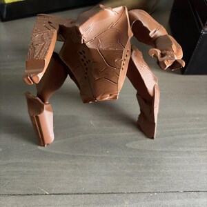 May include: A brown, multi-legged robotic figurine with a segmented, angular body and textured surface details. The object features a central torso with visible layered lines from 3D printing, multiple articulated limbs, and small perforated holes on the front panel.