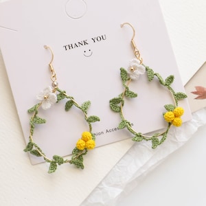 May include: A pair of gold hoop earrings with crocheted green leaves and white and yellow flowers. The earrings have a "Thank You" card behind them.