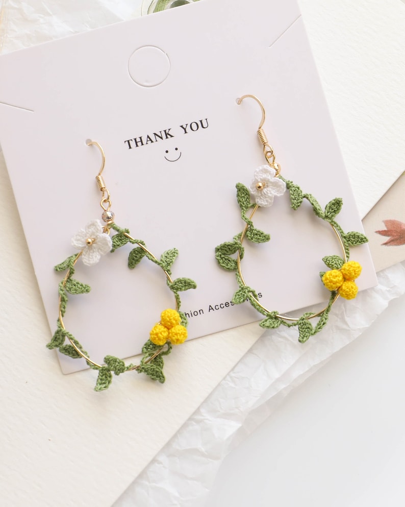 May include: A pair of gold hoop earrings with crocheted green leaves and white and yellow flowers. The earrings have a "Thank You" card behind them.