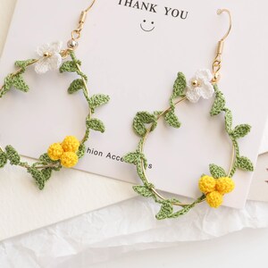 May include: A pair of gold hoop earrings with crocheted white flowers and yellow flower clusters, green leaves, and a gold hook.