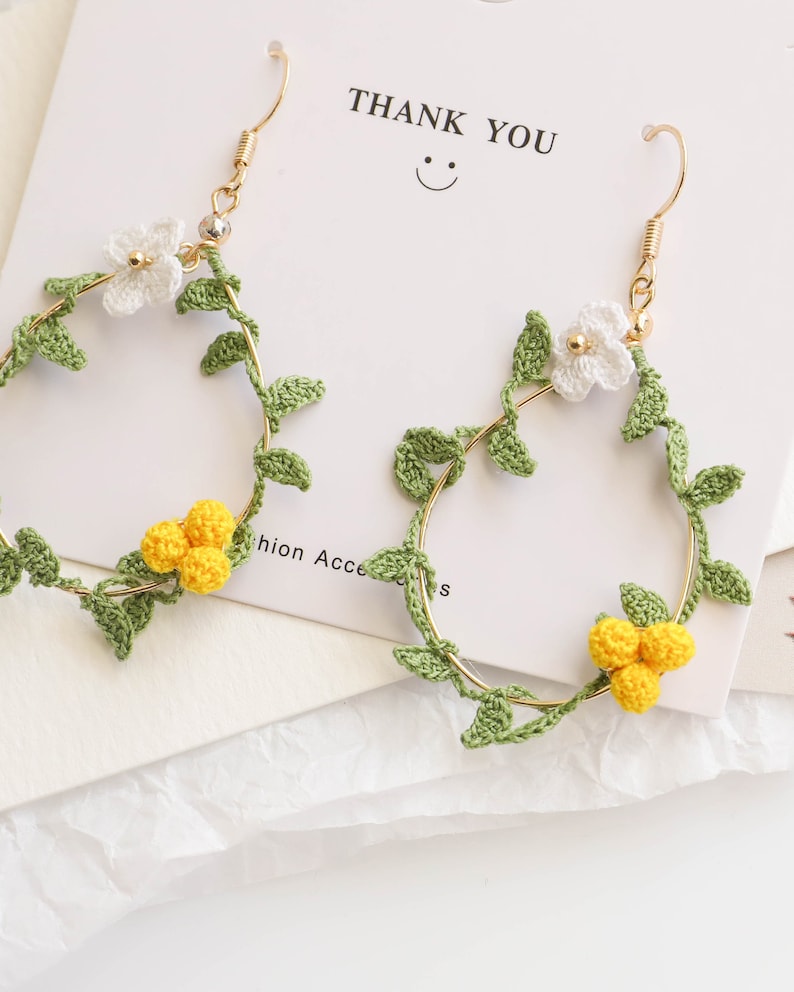 May include: A pair of gold hoop earrings with crocheted white flowers and yellow flower clusters, green leaves, and a gold hook.