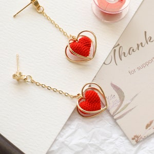 May include: A pair of gold dangle earrings with a red crocheted heart charm inside a gold heart frame. The earrings have a simple gold post and chain.