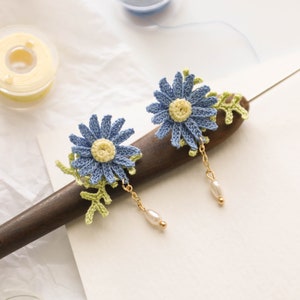 May include: A pair of crocheted flower earrings with blue and green flowers and a pearl dangle. The earrings are displayed on a wooden surface.